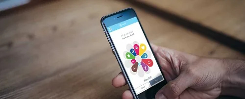 Hand holding a smartphone displaying a colorful pinwheel app interface with multiple icons, resting on a wooden surface.